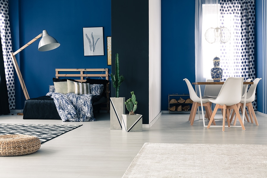 A modern living space blending a bedroom and dining area in a contemporary design. The bedroom features a wooden bed frame with blue and white bedding, complemented by a sleek floor lamp and decorative pillows. The dining area includes a wooden table with white chairs, a blue-and-white vase centerpiece, and a unique chandelier. The space is accented with potted cacti, patterned curtains, and framed artwork, creating a cozy and stylish ambiance.