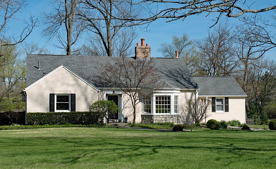 Image of a beautifully painted single-story house with light beige exterior and dark shutters, showcasing professional exterior painting services. The home features a well-maintained lawn, mature trees, and a clean, inviting appearance, highlighting the quality and attention to detail in the painting work.