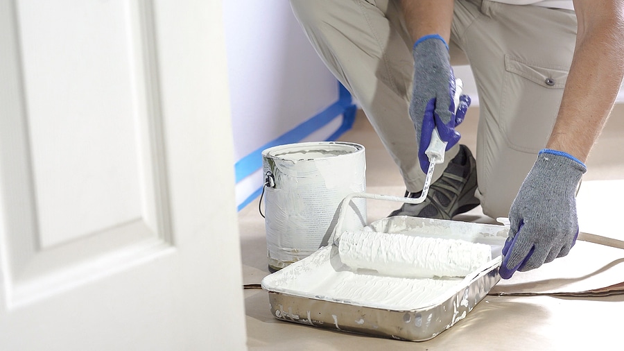 Professional painter wearing gray gloves and beige work pants prepares a paint roller with white paint from a metal tray, with a white paint can nearby and blue painter’s tape along the wall baseboard; protective floor covering in place for a clean and mess-free interior painting job.